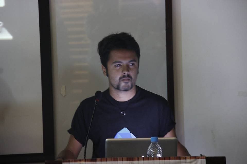 Fahad standing at a lectern behind a laptop, mid-presentation in a darkened room.