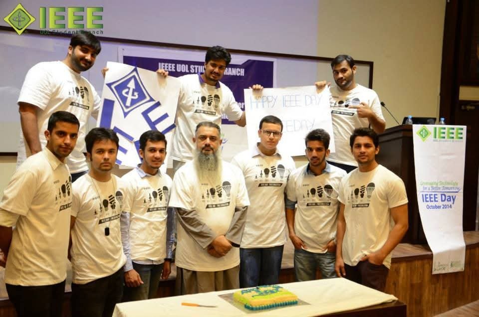 Group photo of the IEEE UOL Student Branch team holding handmade IEEE Day signs, with a cake on the table in front.