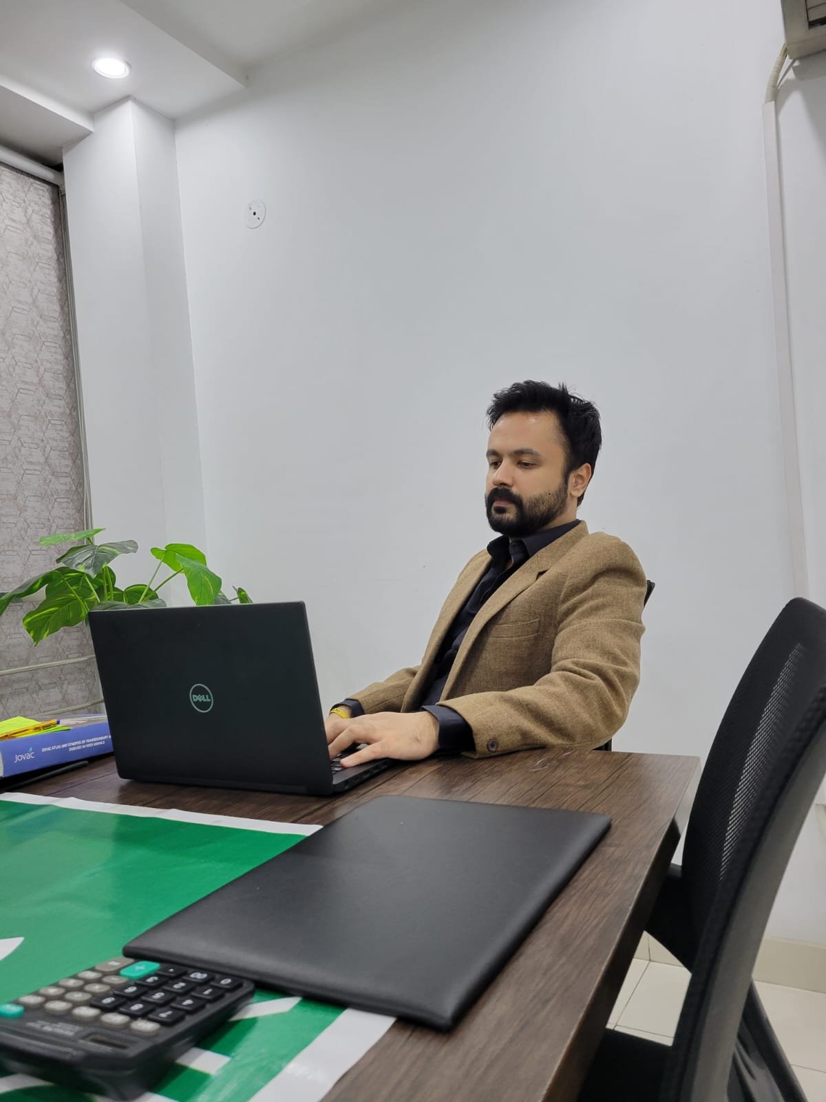 Fahad working at a laptop at an office desk, wearing a tan blazer over a dark shirt.